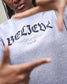 Person wearing a gray sweatshirt with 'BELIEVE' printed on it, pointing at the text.