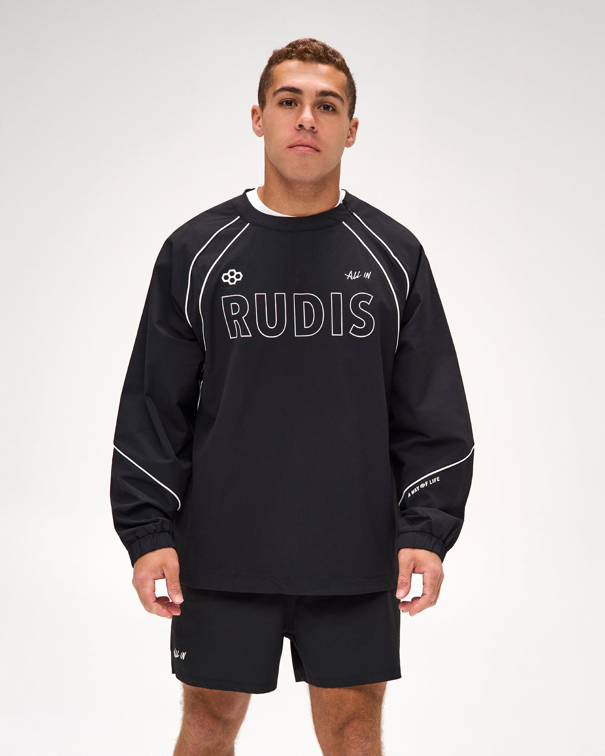 A man modeling a black long-sleeve athletic sweatshirt with white piping and 'RUDIS' printed across the chest, paired with matching black shorts against a light gray studio background.