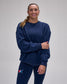 A woman wearing a navy blue crewneck sweatshirt and matching navy blue leggings with a small red, white, and blue logo on the left thigh, standing against a light gray studio background.