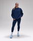 A woman modeling a matching navy blue activewear set consisting of a crewneck sweatshirt and high-waisted leggings, paired with white socks and light blue sneakers, against a neutral gray studio background.