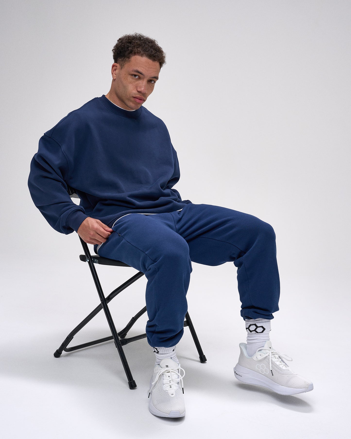 Man sitting on a chair wearing a navy tracksuit and white sneakers against a white background