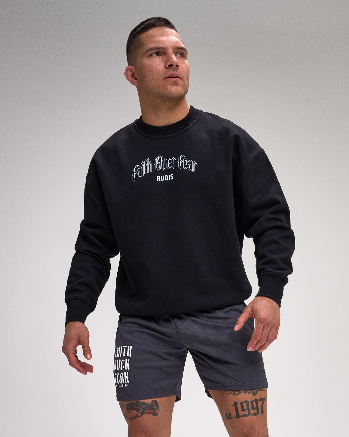 Man wearing a black sweatshirt with text and gray shorts on a plain background