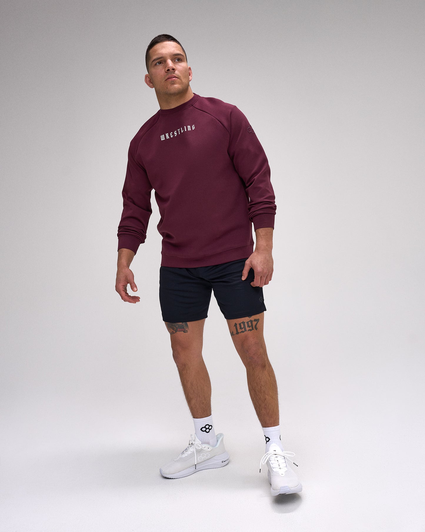 Man wearing a maroon sweatshirt with text, black shorts, white socks, and sneakers on a gray background