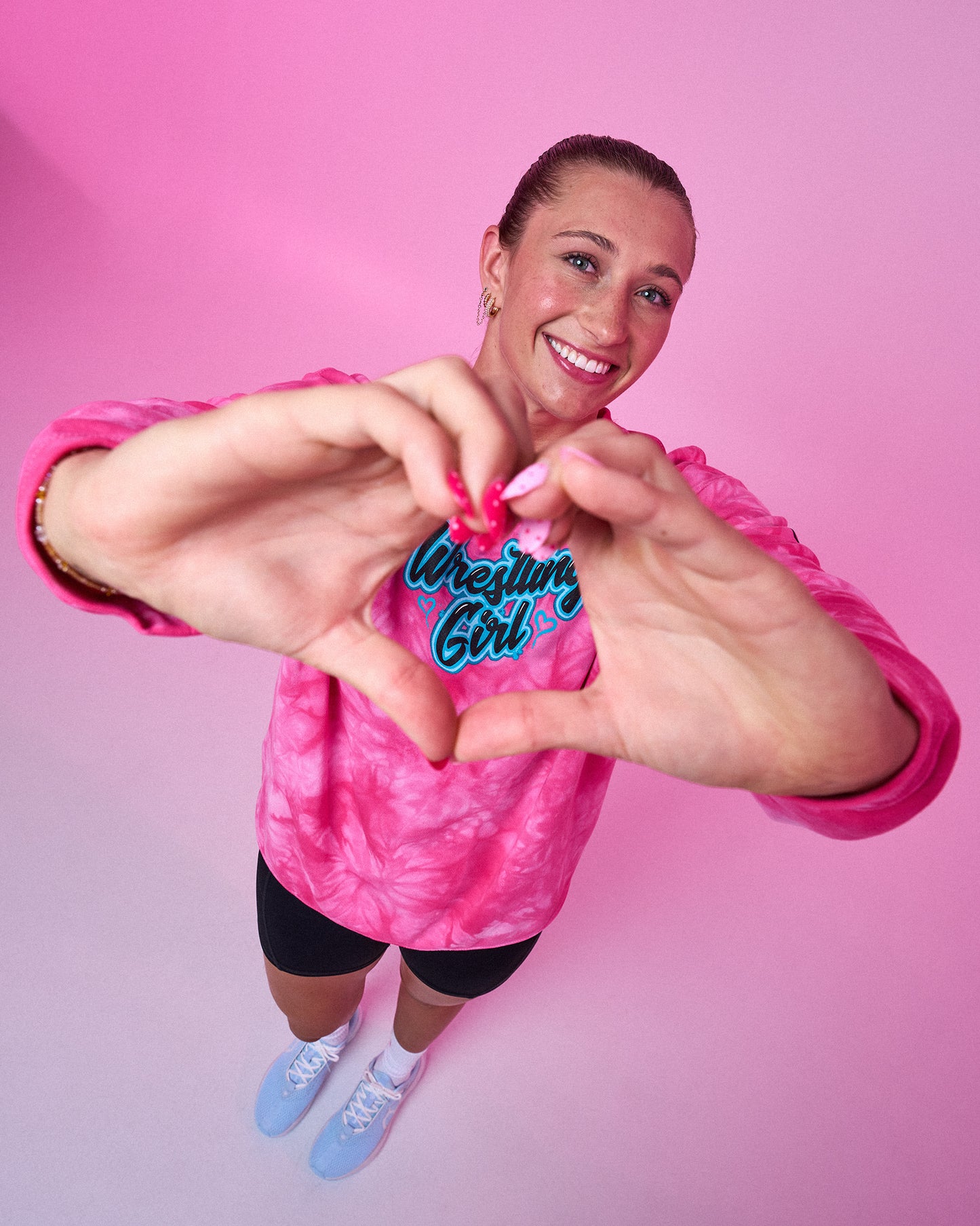 A smiling woman wearing a pink tie-dye long-sleeve shirt with 'Wrestling Girl' printed in blue and white on the chest, posing with her hands forming a heart shape over the text. She is standing against a solid pink background.