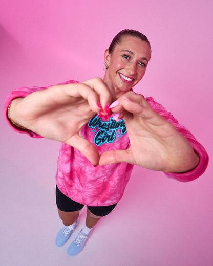A smiling woman wearing a pink tie-dye long-sleeve shirt with 'Wrestling Girl' printed in blue and white on the chest, posing with her hands forming a heart shape over the text. She is standing against a solid pink background.