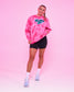 A woman modeling a pink tie-dye sweatshirt with 'Wrestling Girl' printed in teal across the chest, paired with black athletic shorts, white socks, and light purple sneakers, set against a solid pink studio background.