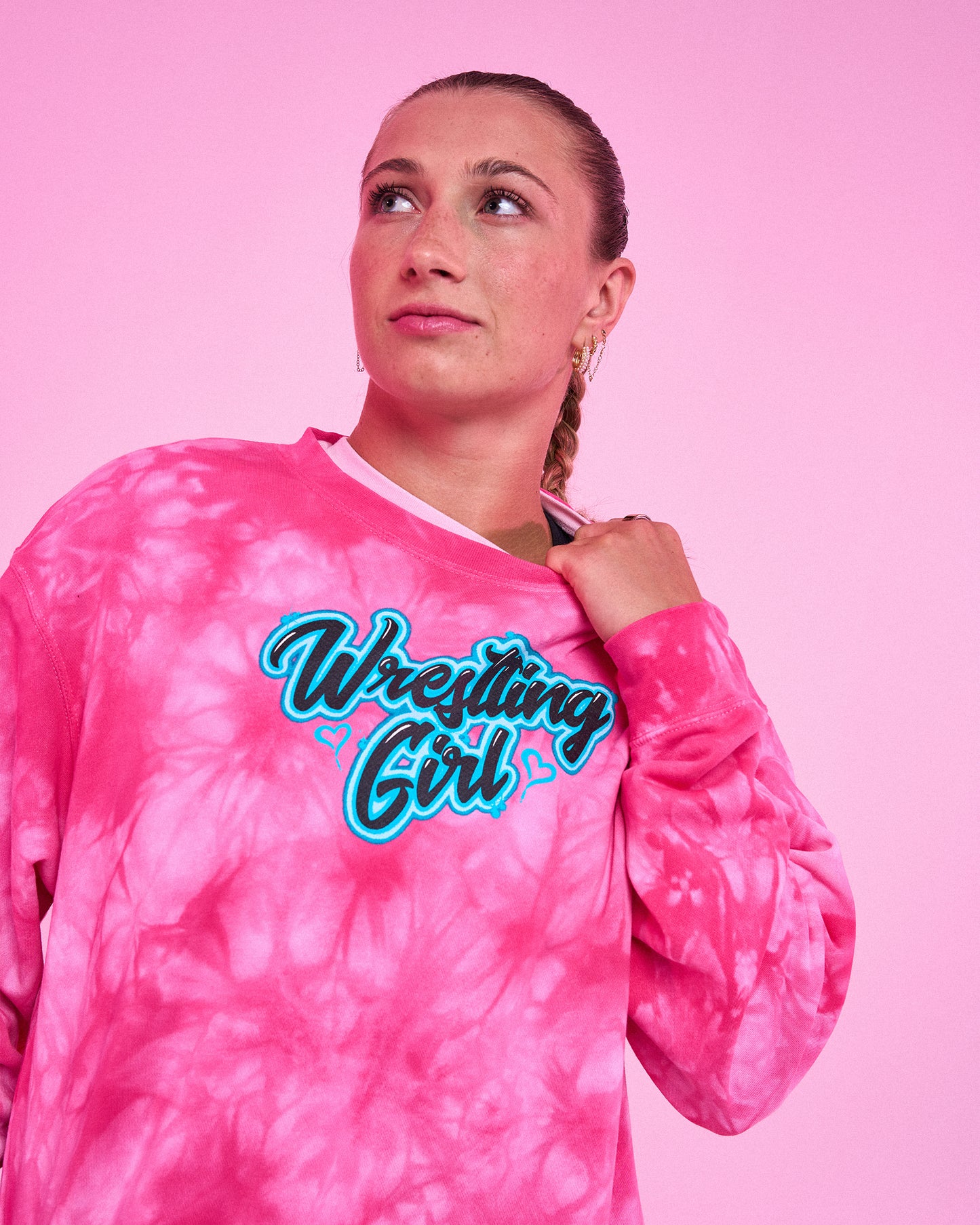 A woman wearing a pink tie-dye long-sleeve sweatshirt with 'Wrestling Girl' printed in a stylized blue and white graphic across the chest, posed against a solid pink background.