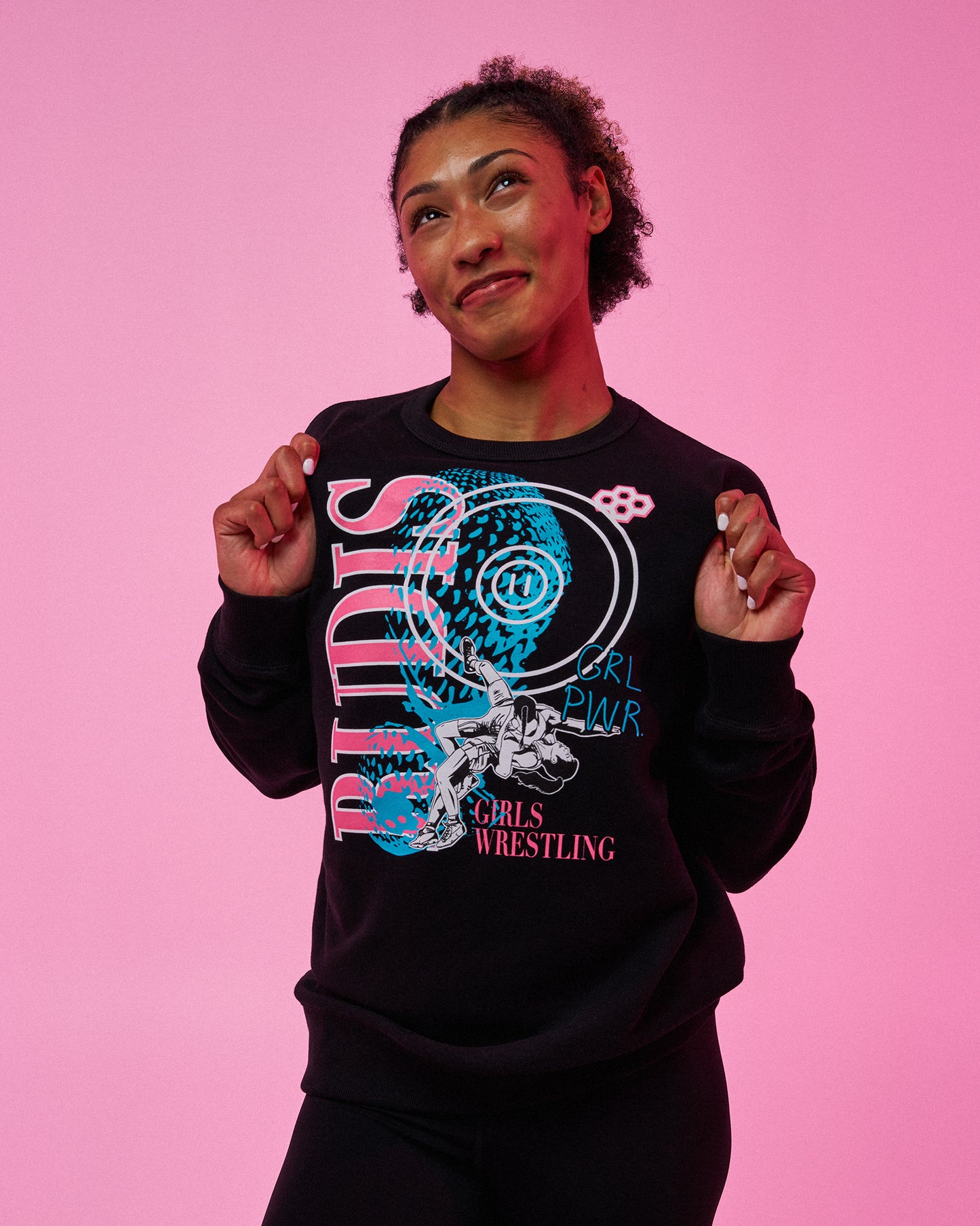 A woman posing against a solid pink background wearing a black crewneck sweatshirt with a colorful graphic that reads 'RUDIS GIRL Power GIRLS WRESTLING'.