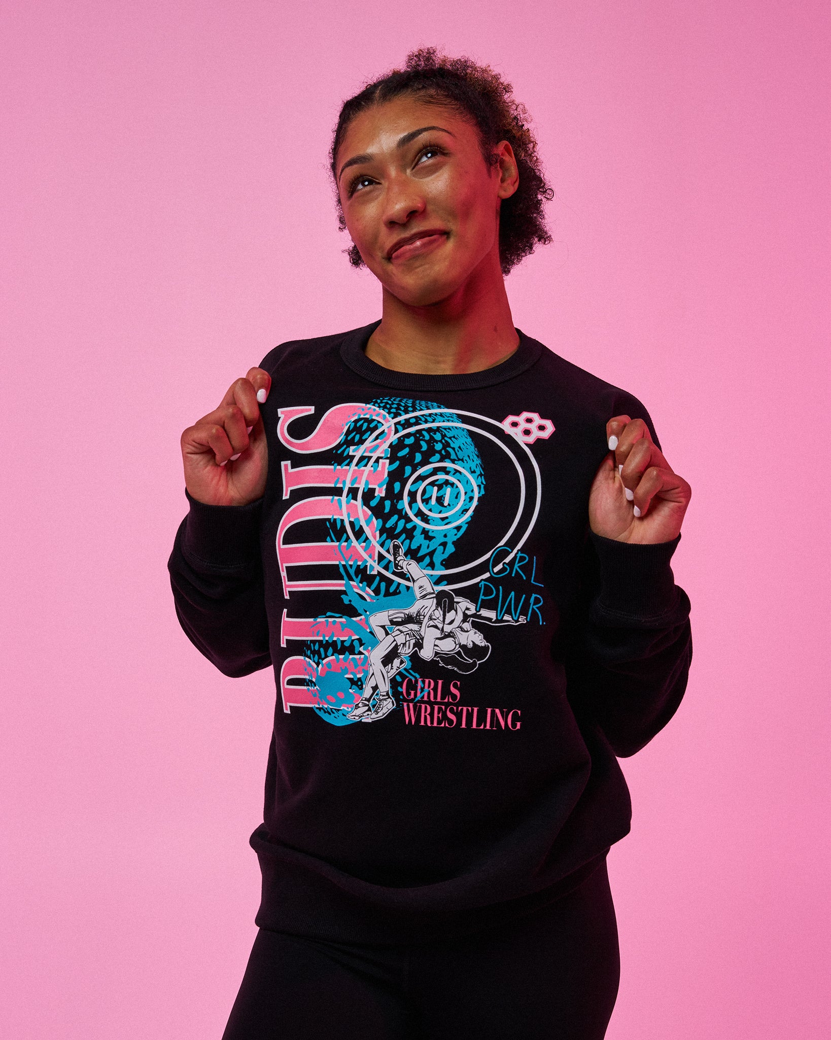 A woman posing against a solid pink background wearing a black crewneck sweatshirt with a colorful graphic that reads 'RUDIS GIRL Power GIRLS WRESTLING'.