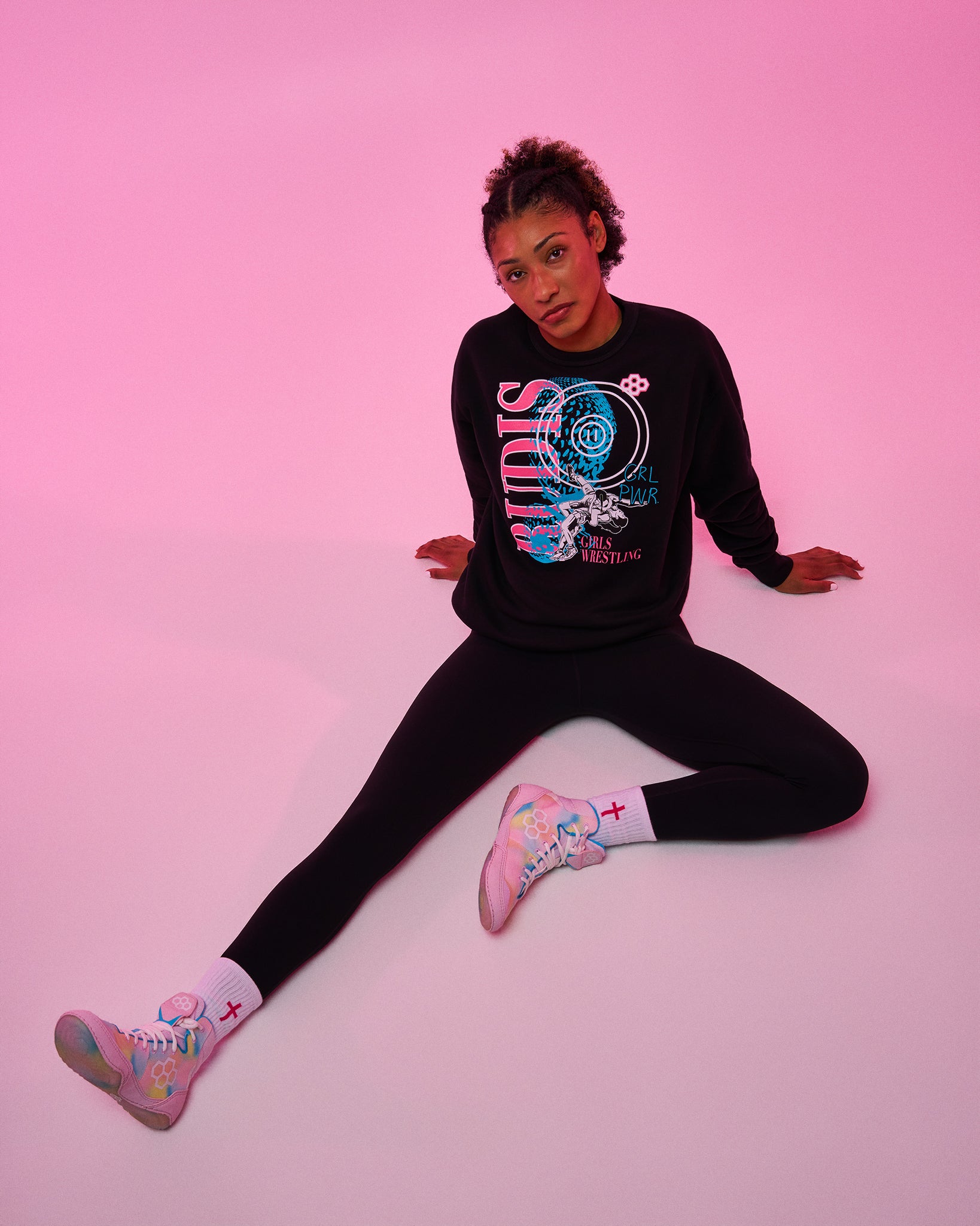A model sits on a pink studio background wearing a black crewneck sweatshirt with a colorful graphic print, black leggings, and pastel pink sneakers with white socks.
