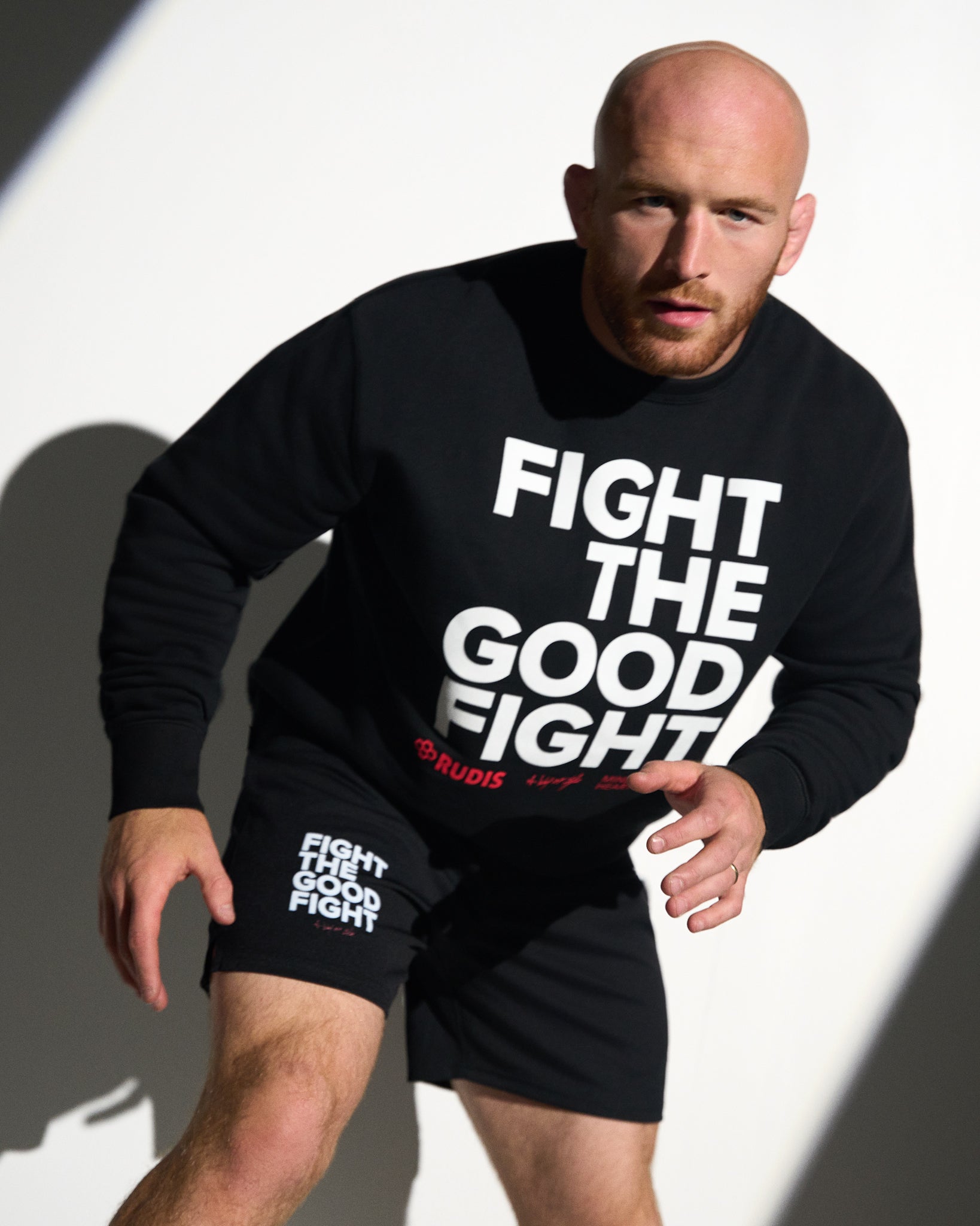 Athlete wearing the RUDIS Fight the Good Fight Comfort Fleece Crewneck in black with matching shorts, posed in a wrestling stance to showcase the bold graphic design and premium athletic fit.