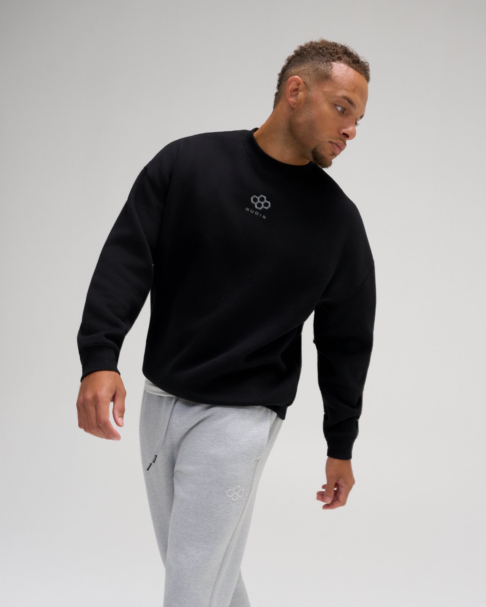 Man wearing the RUDIS Hex Relaxed Comfort Fleece Crewneck in Black with a tonal chest logo, relaxed silhouette, and ribbed cuffs.
 Copy alt text