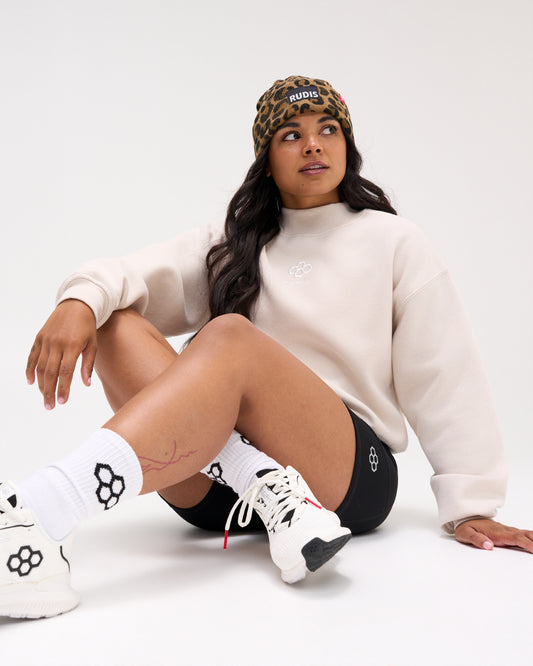 Woman seated wearing the RUDIS Hex Comfort Fleece Mockneck in Bone with black athletic shorts, leopard-print beanie, and white wrestling shoes.
 Copy Alt Text