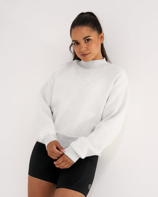 Woman wearing the RUDIS Hex Comfort Fleece Mockneck in Heather Silver with black athletic shorts, showing the relaxed silhouette and subtle chest logo.
Copy Alt Text