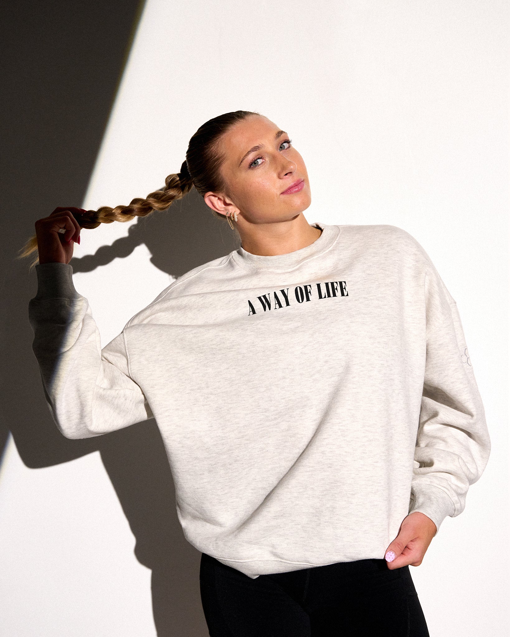A woman wearing a light heather gray sweatshirt with the black text 'A WAY OF LIFE' printed across the chest, posed against a stark white background with dramatic shadows.
