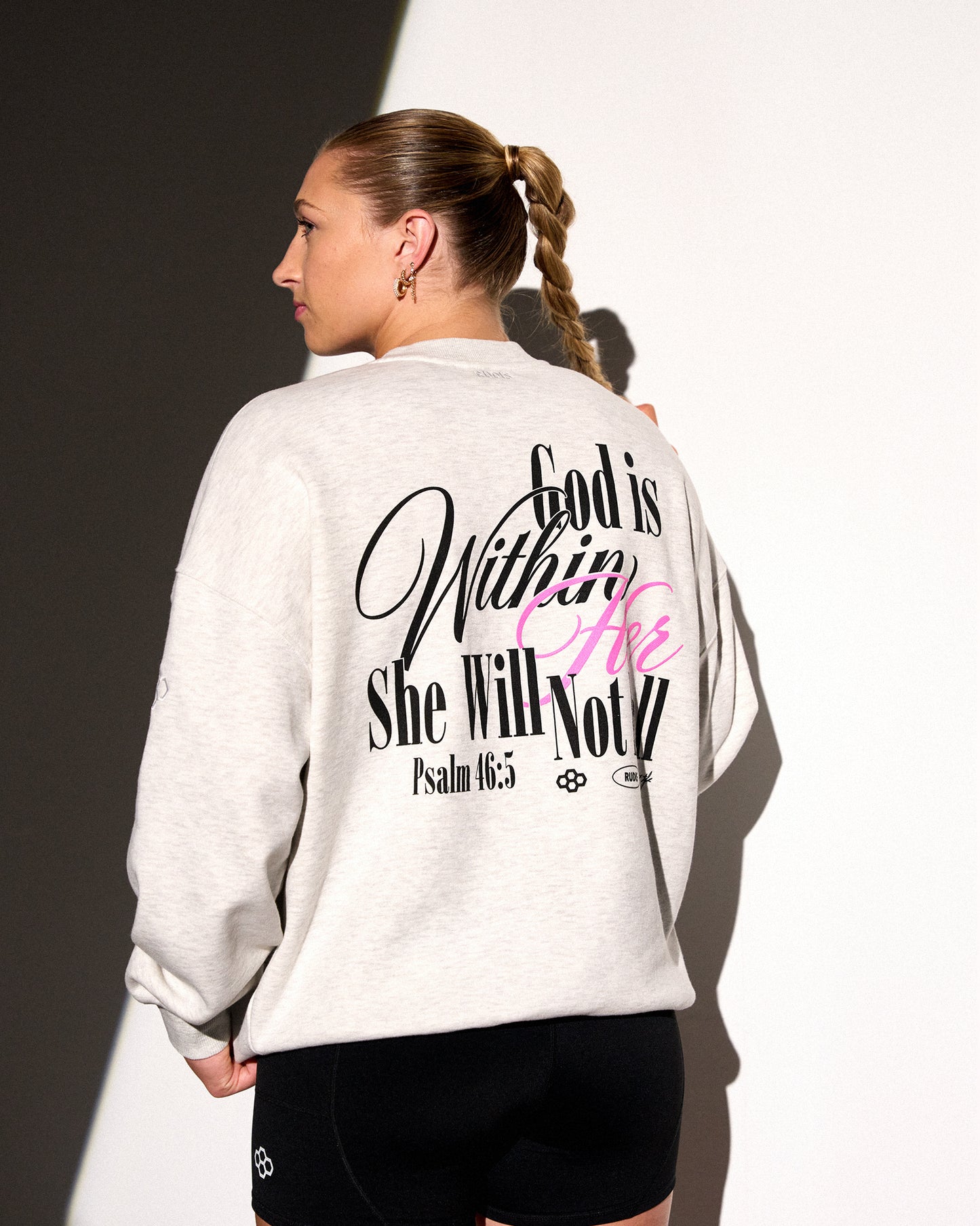A woman shown from the back wearing a light heather gray sweatshirt with a large black and pink Bible verse graphic on the back, paired with black shorts against a white background with a dark shadow.