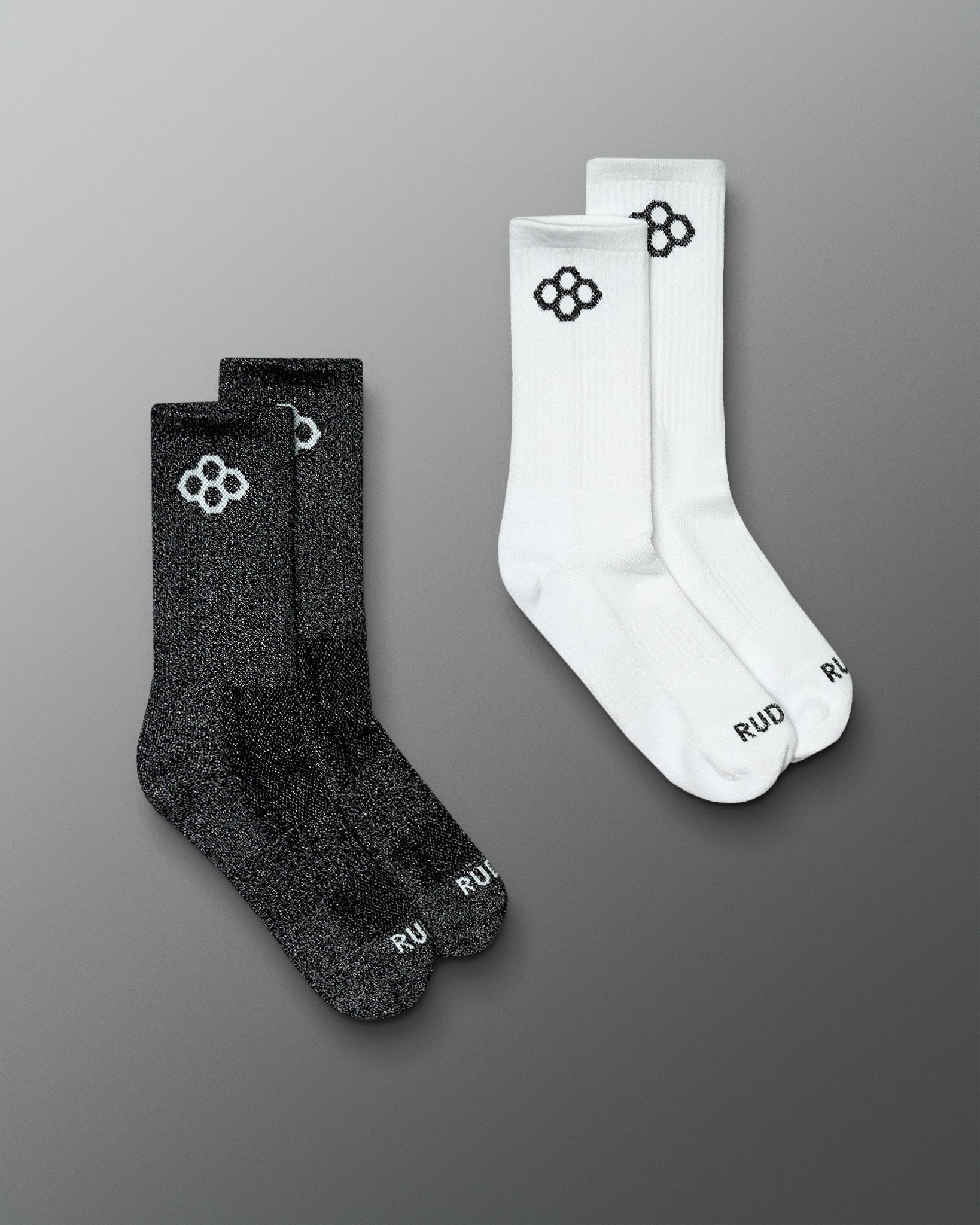 Two pairs of crew socks on a smooth gray background: one pair is black with a white logo and the other pair is white with a black logo. The word 'RUD' is visible near the toes.