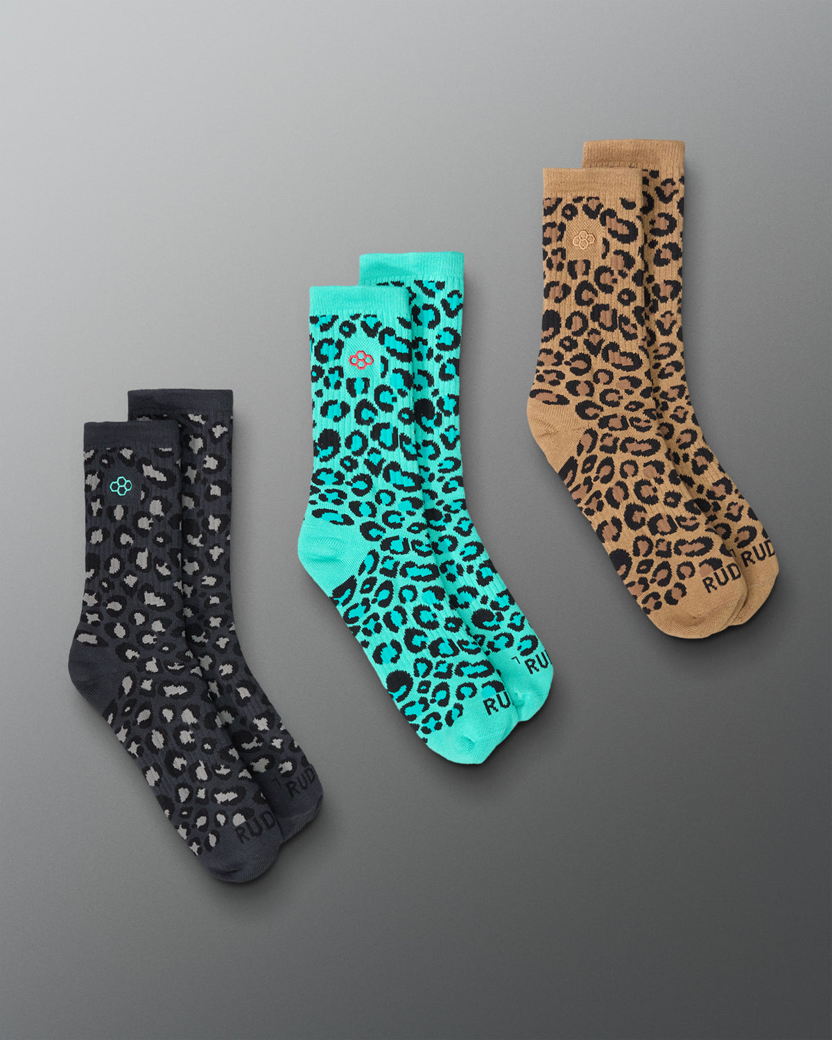 Three pairs of crew socks with animal print patterns arranged diagonally on a gray gradient background. The left pair is dark gray with white spots, the middle pair is turquoise with black leopard spots, and the right pair is tan with black leopard spots.