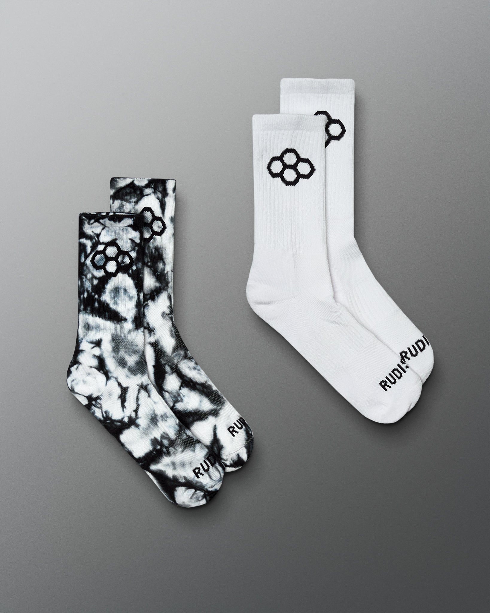 Two pairs of crew socks on a smooth gray gradient background. One pair is white with a black tie dye pattern and black logo, and the other pair is black and white tie-dye with a black logo.