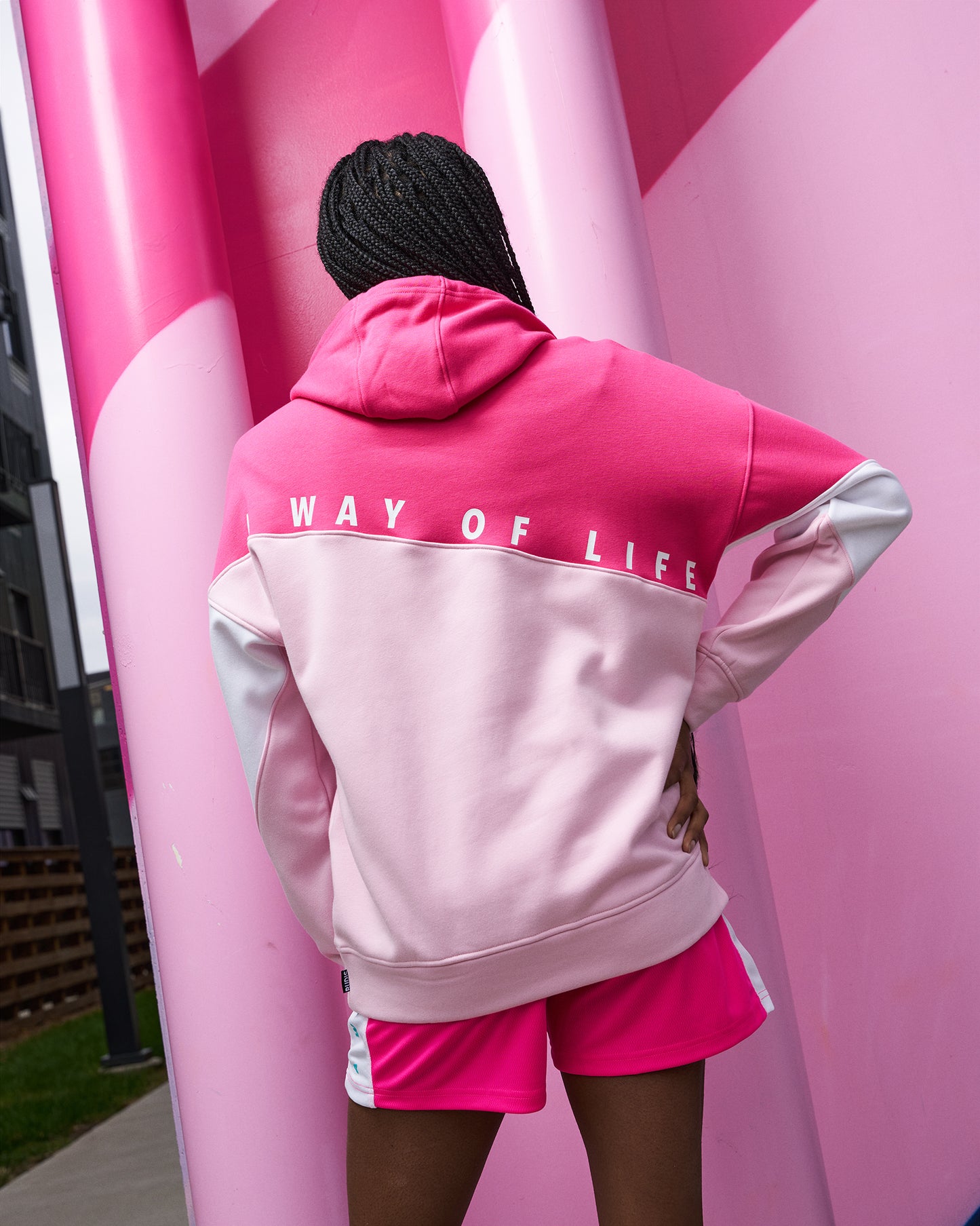 Person wearing a pink 'Way of Life' hoodie and shorts against a pink inflatable structure.