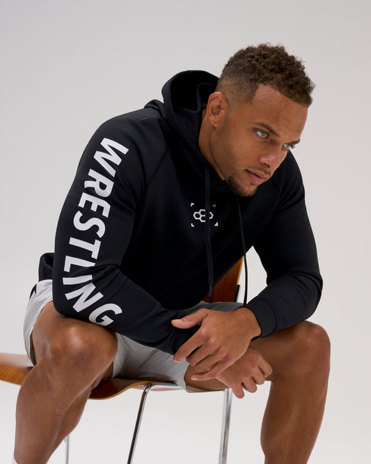 Male athlete wearing the black RUDIS Wrestling COOL-FEEL Hoodie while seated, highlighting the white WRESTLING sleeve graphic and RUDIS chest mark.