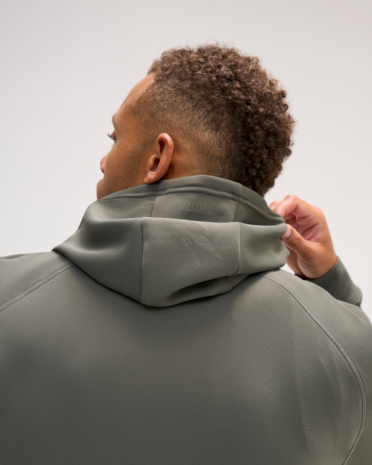 A man shown from behind wearing a dark gray athletic hoodie, adjusting the collar. The brand name 'RUDIS' is subtly printed on the inside of the collar.
