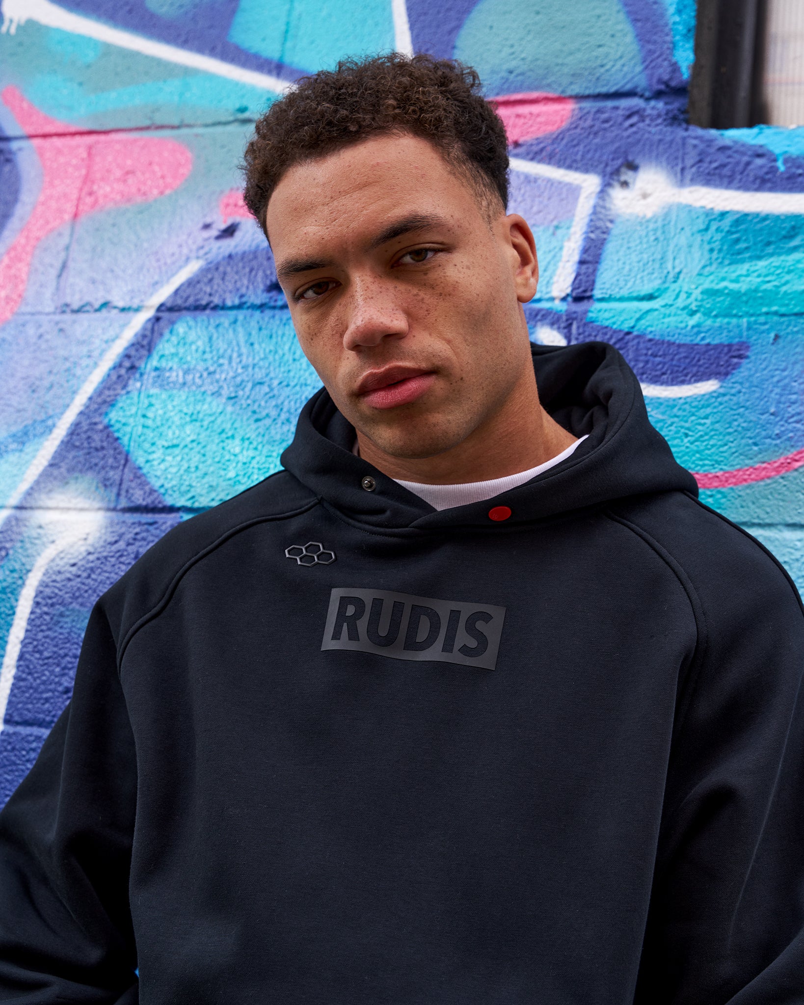 Person wearing a black hoodie with 'RUDIS' logo against a colorful graffiti wall.