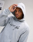 Model wearing the heather gray RUDIS A Way of Life Tech Fleece Hoodie with the hood up, showcasing the black script chest graphic and RUDIS mark.