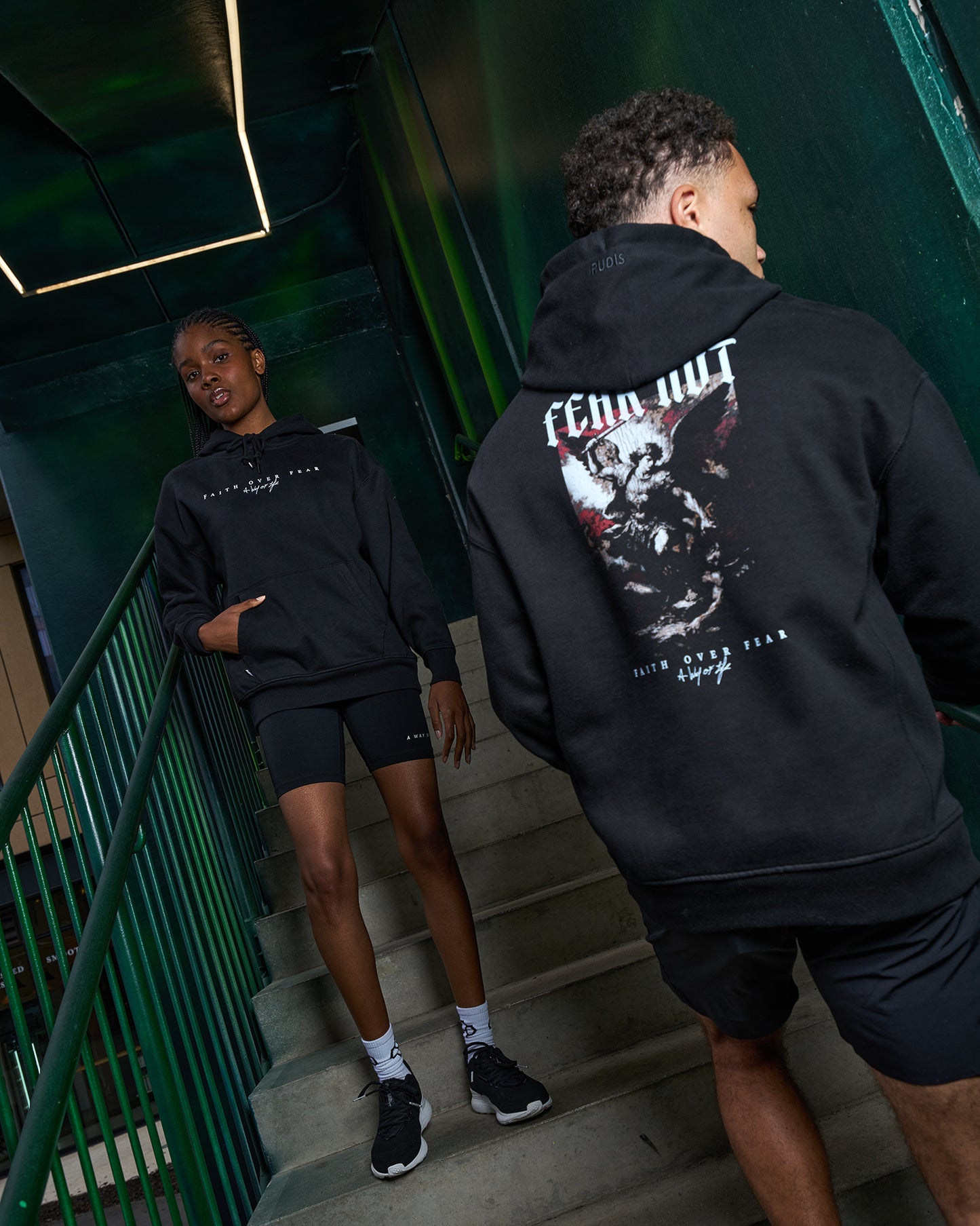 Two people wearing black hoodies with graphic designs on a staircase.