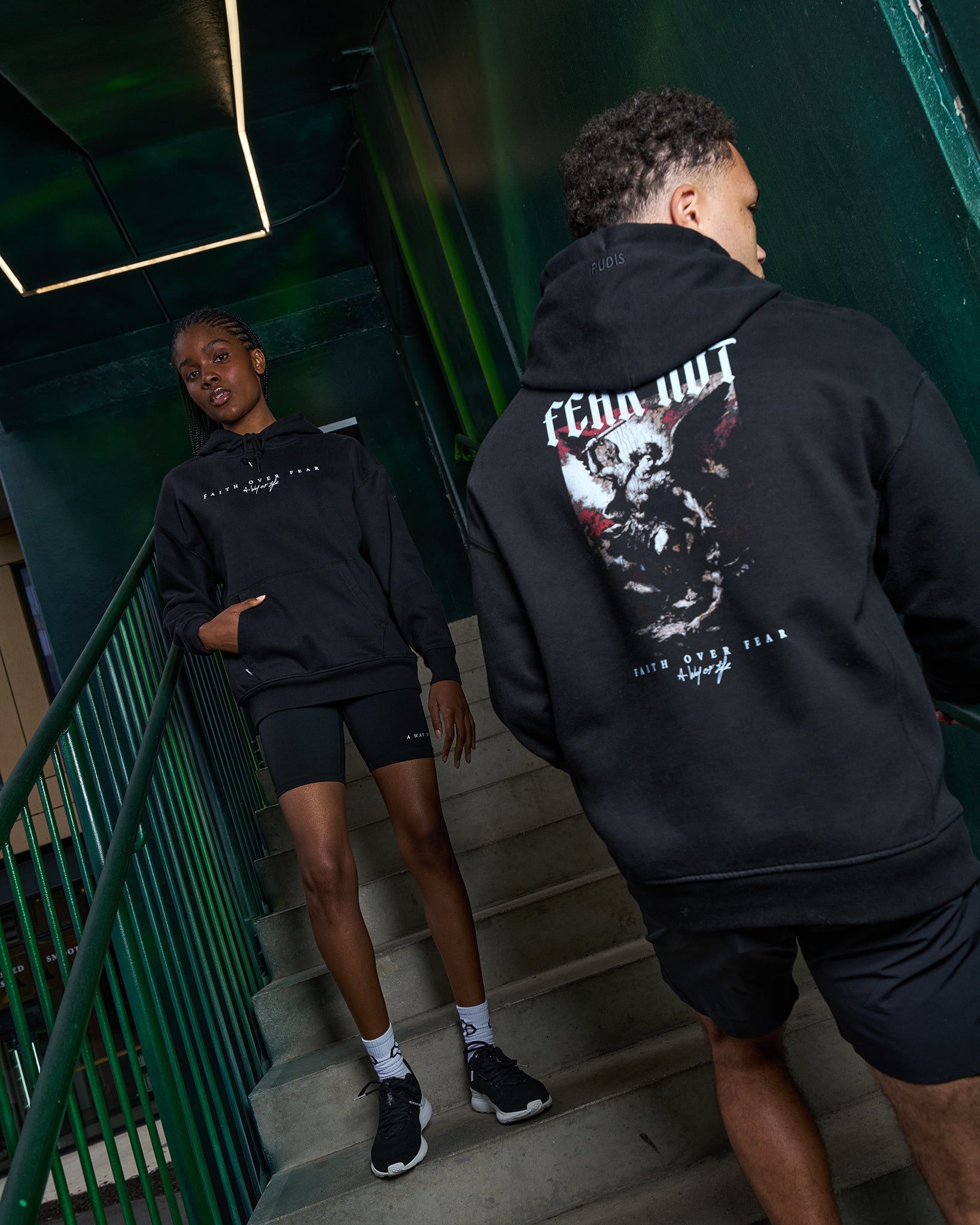 Two people wearing black hoodies with graphic designs on a staircase.