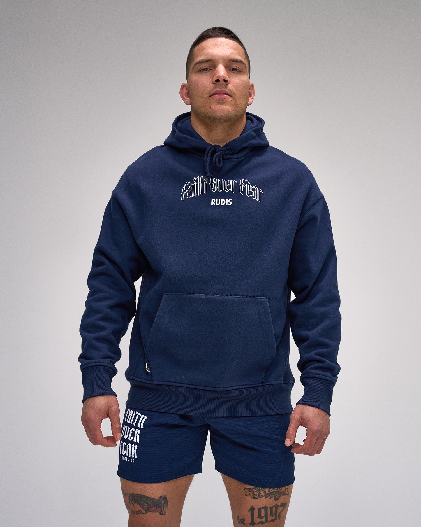 Man wearing a navy hoodie and shorts with 'RUDIS' branding on a gray background