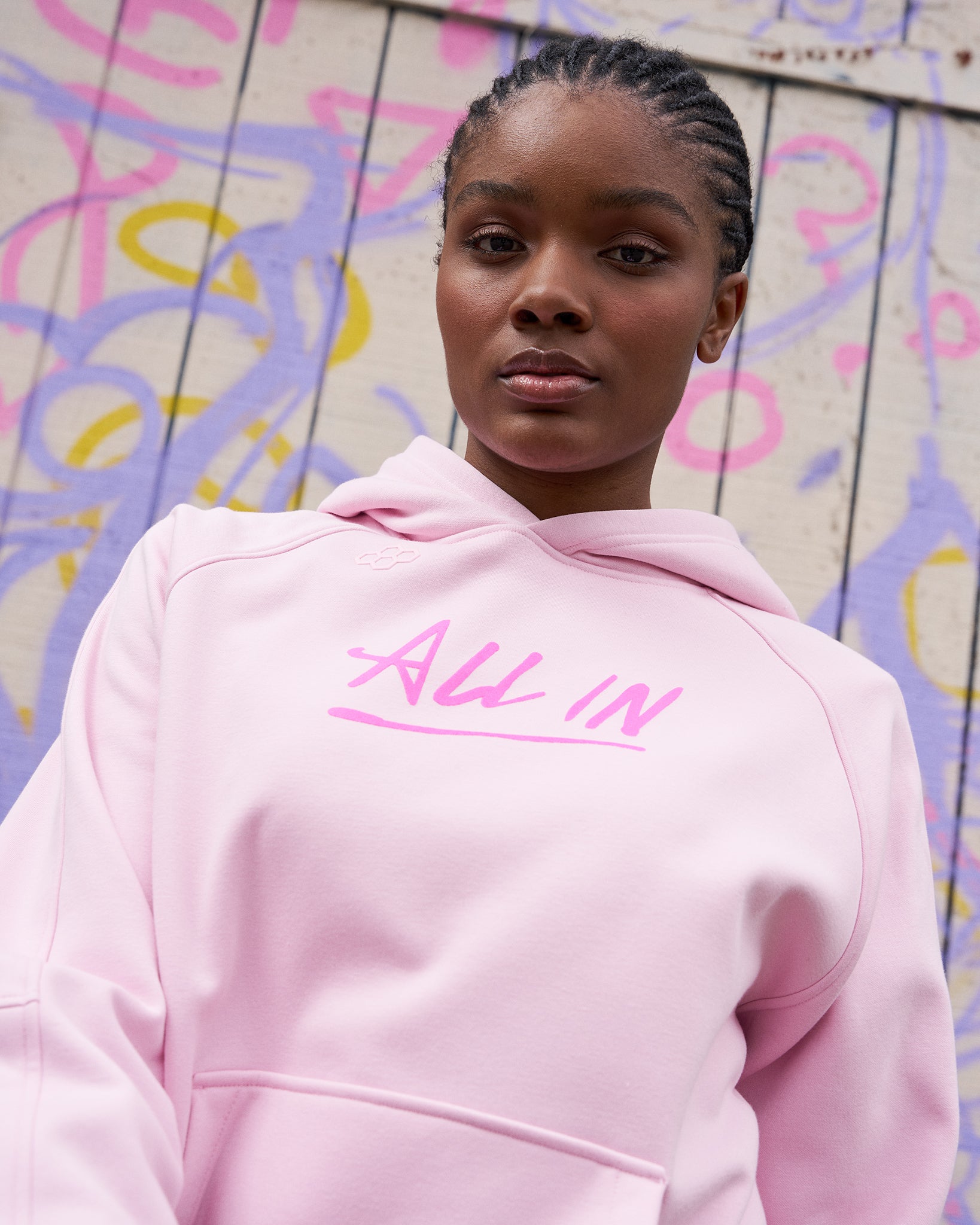 Person wearing a pink hoodie with 'All In' text against a colorful graffiti wall.
