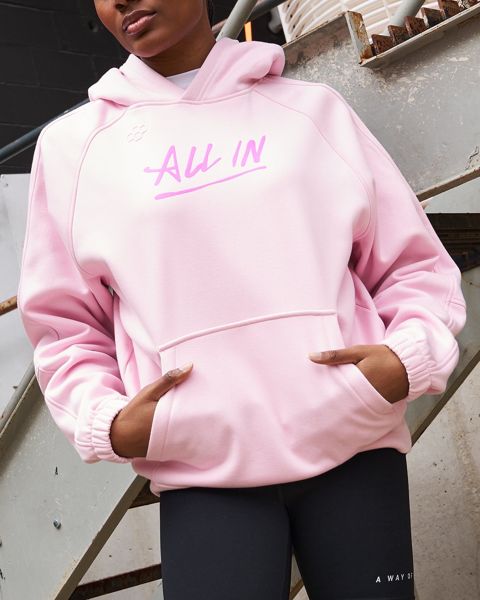 Person wearing a pink hoodie with 'All In' text, standing against a metal railing.