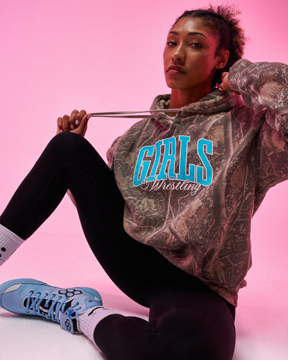 A woman sits against a pink studio background wearing a camouflage-patterned hoodie with 'GIRLS Wrestling' printed in bright blue and white across the chest, paired with black leggings and light blue sneakers.
