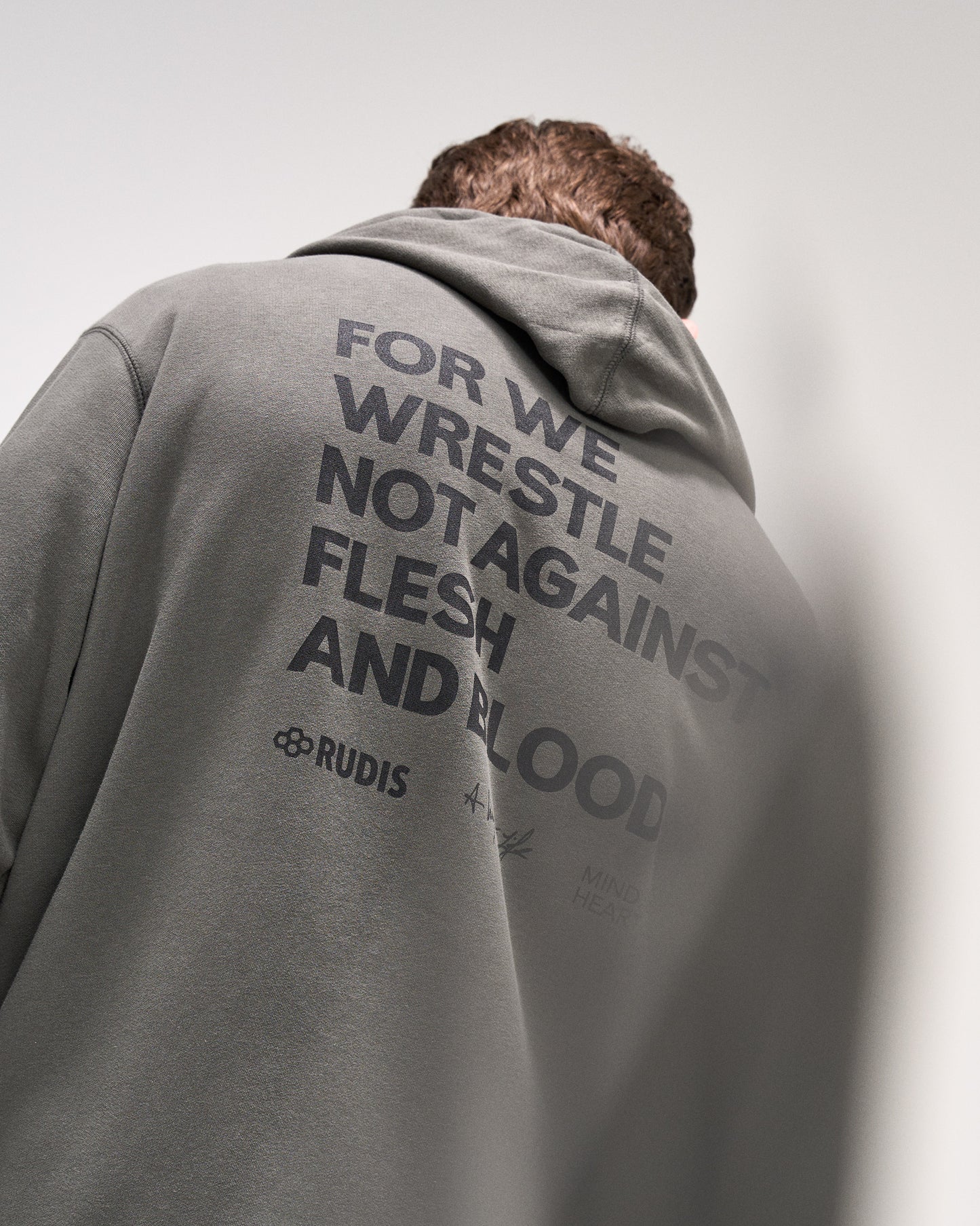 Person wearing a dark olive hoodie with 'For we wrestle not against flesh and blood' text and a brand logo on the back against a plain background