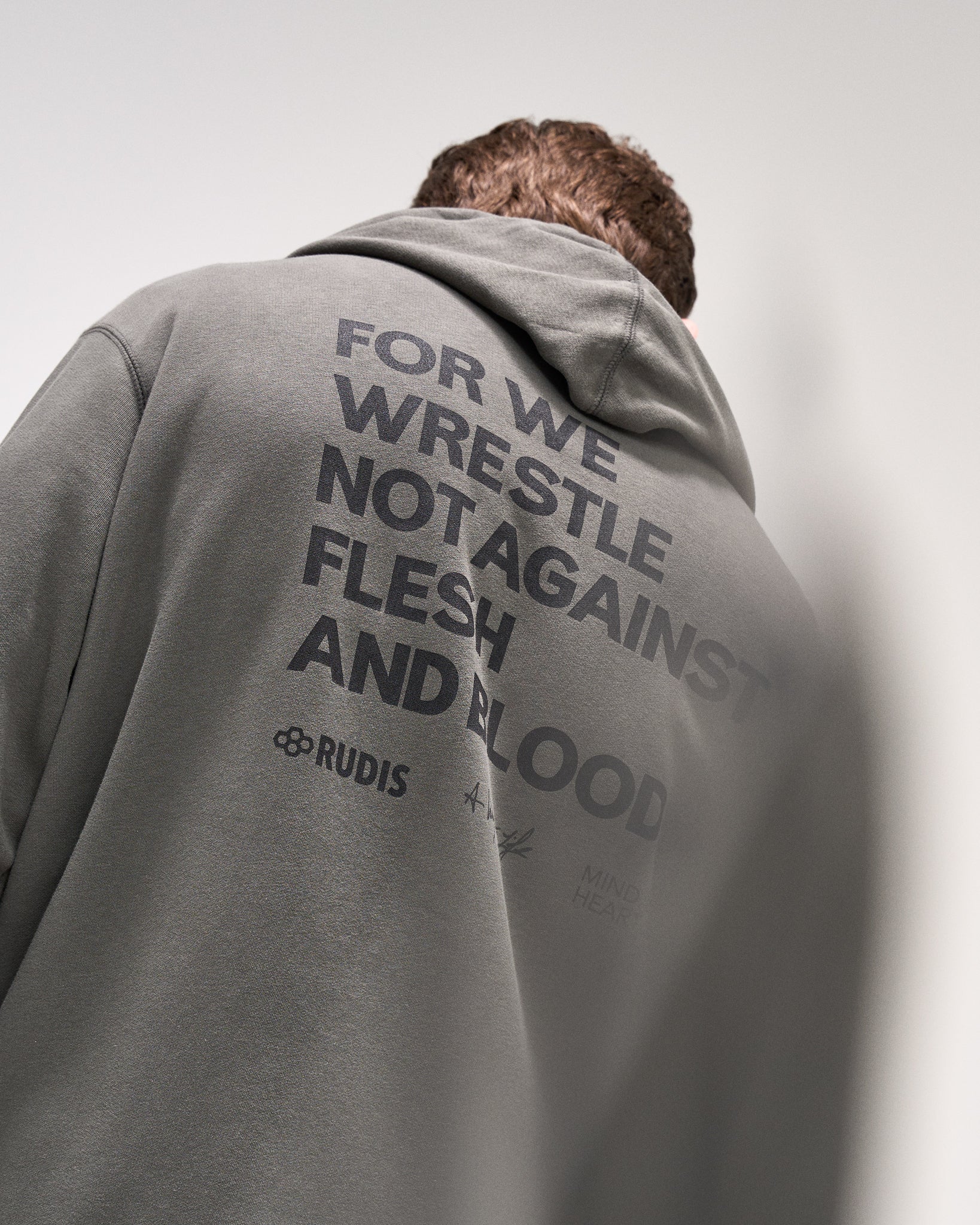 Person wearing a dark olive hoodie with 'For we wrestle not against flesh and blood' text and a brand logo on the back against a plain background