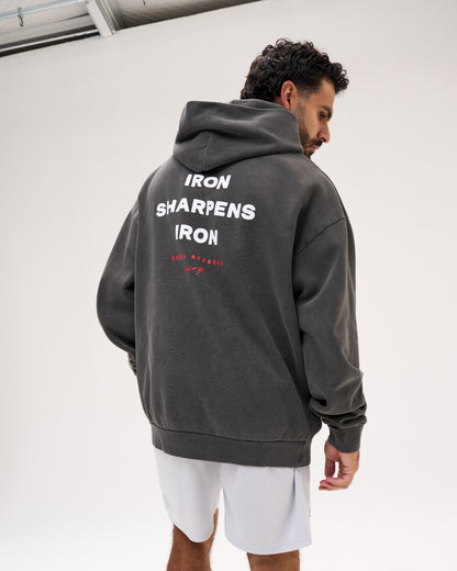 A man wearing a dark gray washed hoodie with 'IRON SHARPENS IRON' printed in white on the back, paired with white shorts, photographed in a studio setting.
