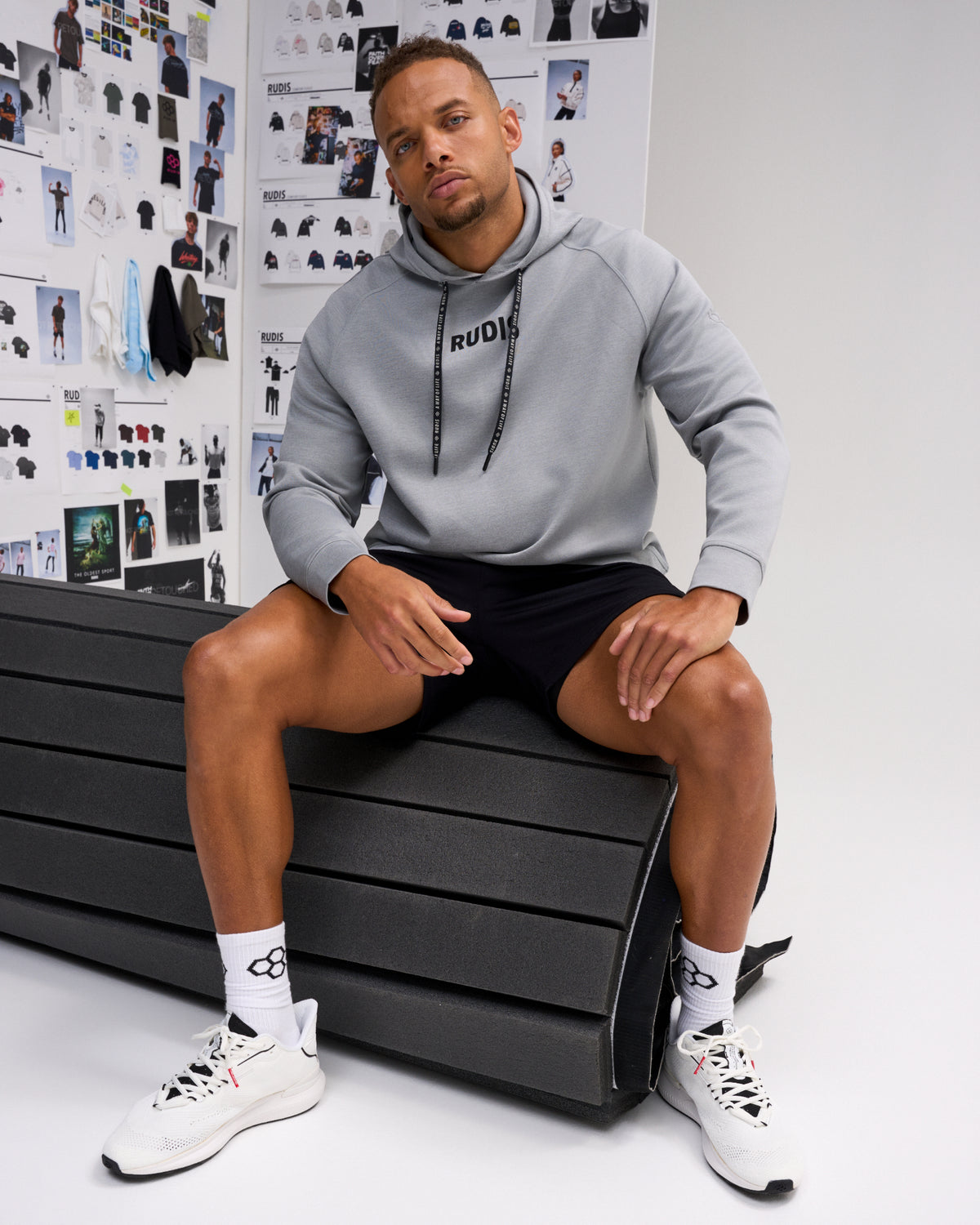 A man wearing a light gray RUDIS hoodie, black athletic shorts, white socks, and white sneakers sits on a black bench in front of a white wall covered with fashion sketches.