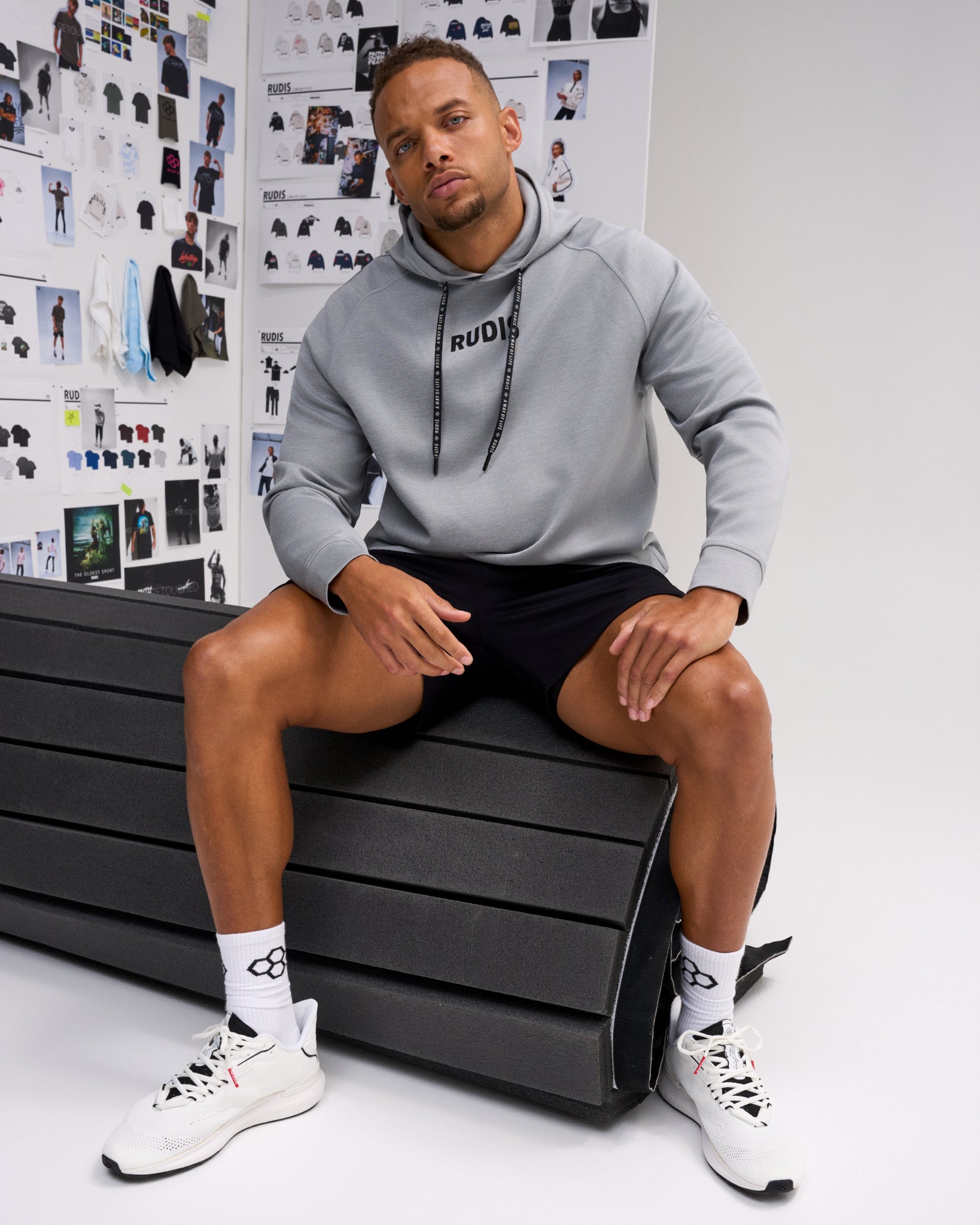 A man wearing a light gray RUDIS hoodie, black athletic shorts, white socks, and white sneakers sits on a black bench in front of a white wall covered with fashion sketches.