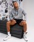 A man wearing a light gray RUDIS hoodie, black athletic shorts, white socks, and white sneakers sits on a black bench in front of a white wall covered with fashion sketches.