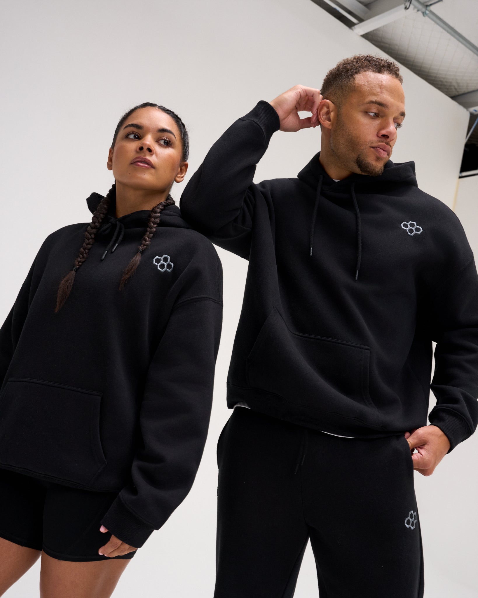 Male and female models wearing the RUDIS Essential Hex Relaxed Comfort Fleece Hoodie in black, featuring a subtle Hex chest logo and kangaroo pockets.