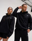 Male and female models wearing the RUDIS Essential Hex Relaxed Comfort Fleece Hoodie in black, featuring a subtle Hex chest logo and kangaroo pockets.
