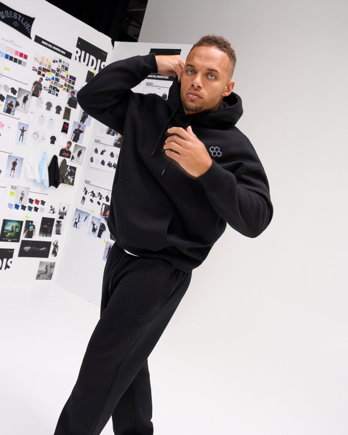 Male model wearing the black RUDIS Essential Hex Relaxed Comfort Fleece Hoodie with matching joggers, showing the relaxed fit and drawstring hood.