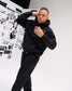 Male model wearing the black RUDIS Essential Hex Relaxed Comfort Fleece Hoodie with matching joggers, showing the relaxed fit and drawstring hood.
