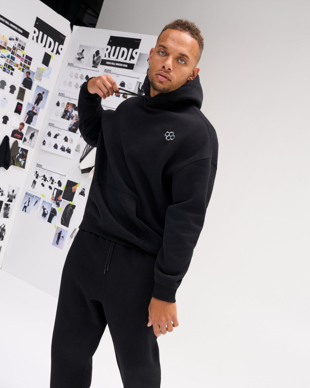Male model styling the RUDIS Essential Hex Relaxed Comfort Fleece Hoodie in black, featuring a minimal chest logo and relaxed silhouette.