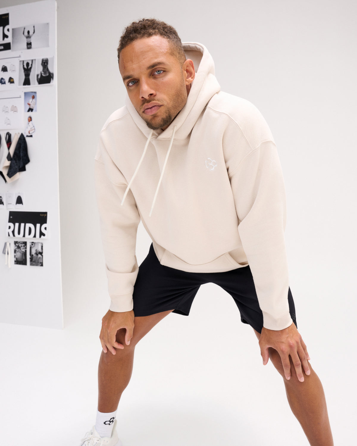 Male model wearing the RUDIS Essential Hex fleece hoodie in Bone with black shorts, showcasing the relaxed fit, tonal chest logo and kangaroo pocket.