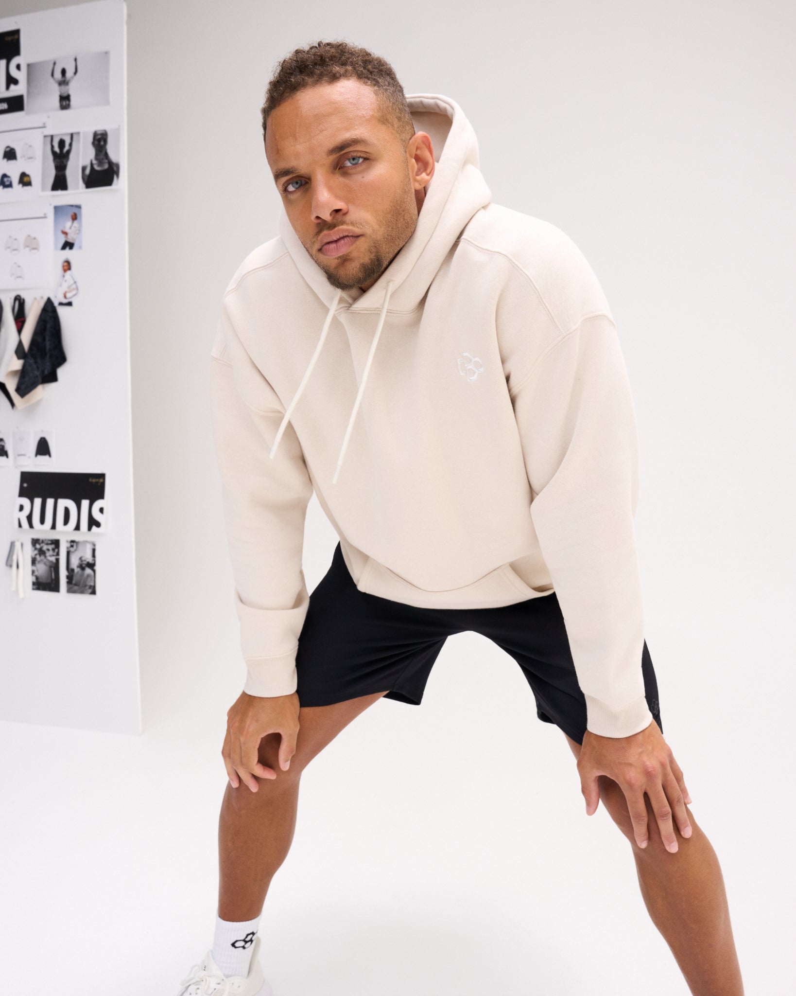 Male model wearing the RUDIS Essential Hex fleece hoodie in Bone with black shorts, showcasing the relaxed fit, tonal chest logo and kangaroo pocket.