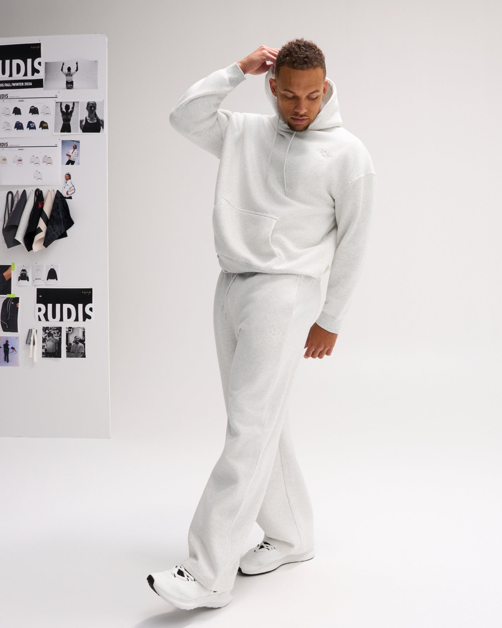 Male model wearing the RUDIS Essential Hex Relaxed Comfort Fleece Hoodie in Heather Silver with matching sweatpants and white athletic shoes.
