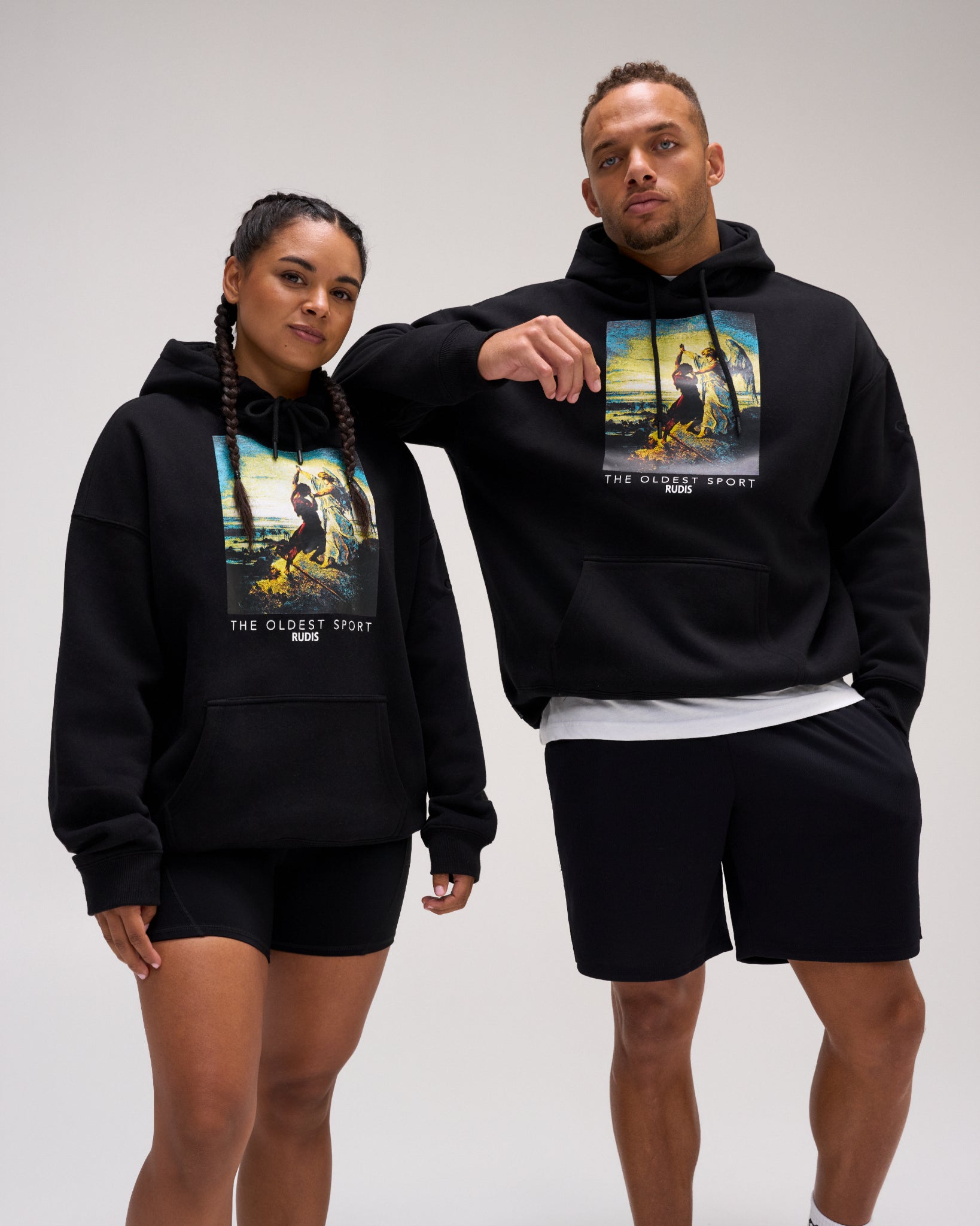 Male and female models wearing the RUDIS Divine Struggle Relaxed Comfort Fleece Hoodie in Black, featuring colorful wrestling-inspired artwork and The Oldest Sport graphic.