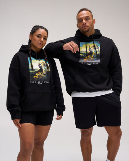 Male and female models wearing the RUDIS Divine Struggle Relaxed Comfort Fleece Hoodie in Black, featuring colorful wrestling-inspired artwork and The Oldest Sport graphic.